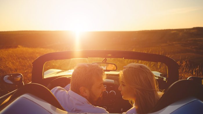 Back view beautiful couple enjoying sunset looking to each other sitting in a sport car while travelling, rays of sun reflecting on them